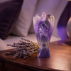 A purple amethyst angel figurine standing on a dark wooden nightstand next to dried lavender sprigs, with a softly blurred bedroom background.