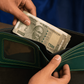A dark green leather bifold wallet with light green contrast stitching, held open by two hands with a stack of Indian 500 Rupee notes being inserted. The wallet features a clear ID window on the front.