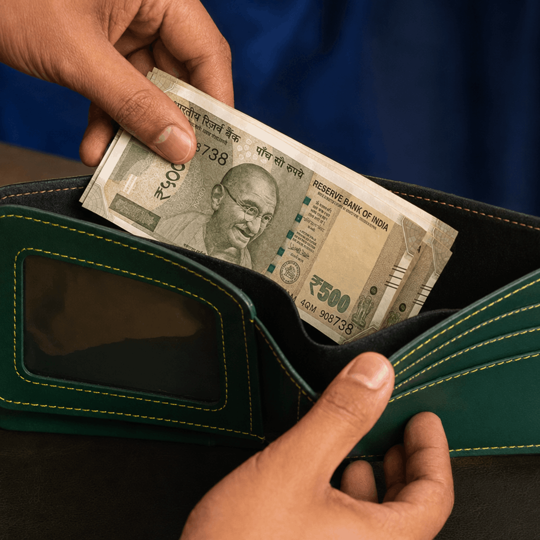 A dark green leather bifold wallet with light green contrast stitching, held open by two hands with a stack of Indian 500 Rupee notes being inserted. The wallet features a clear ID window on the front.
