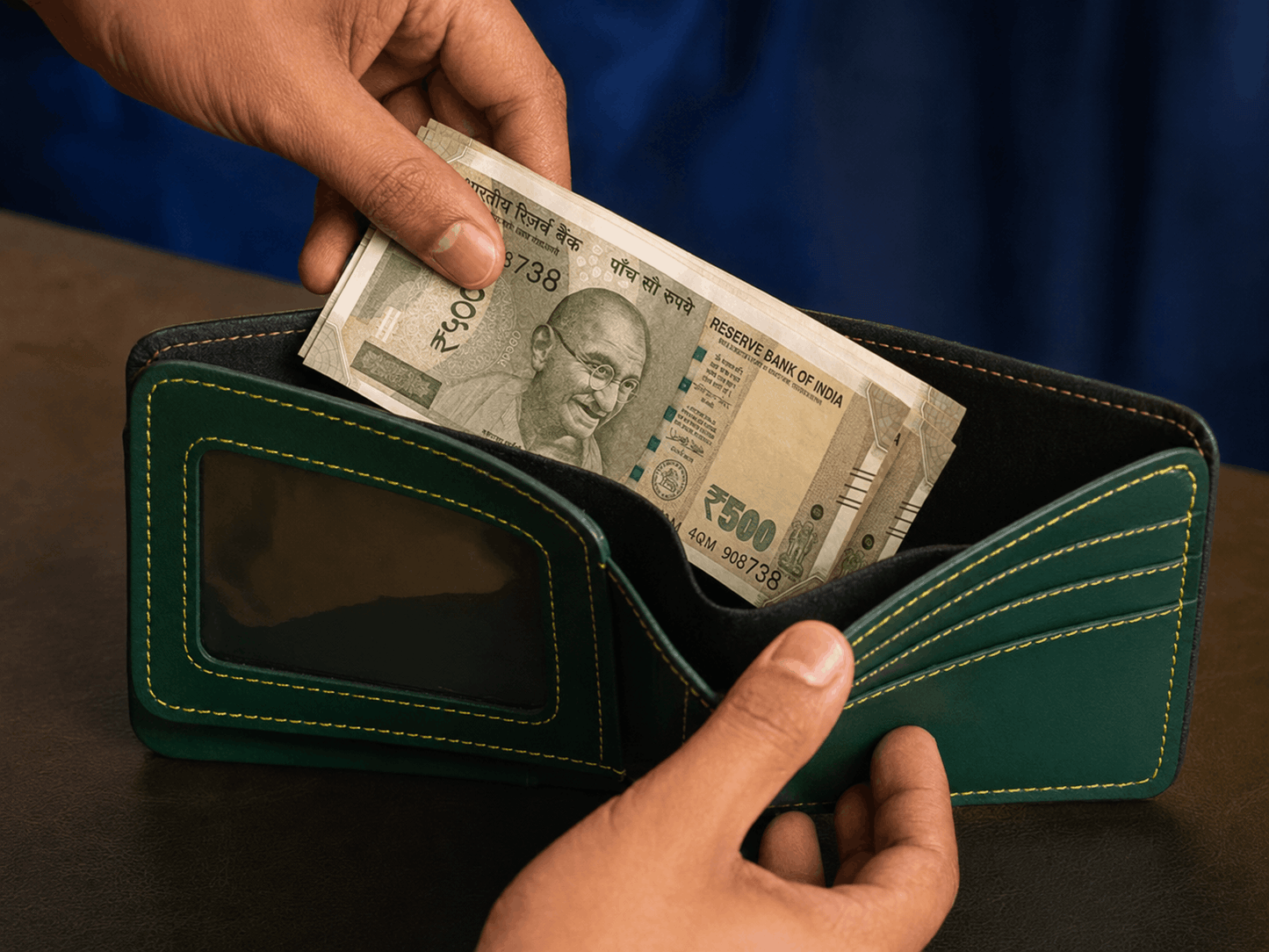 A dark green leather bifold wallet with light green contrast stitching, held open by two hands with a stack of Indian 500 Rupee notes being inserted. The wallet features a clear ID window on the front.
