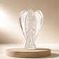 A clear glass angel figurine standing on a round beige stone base against a warm beige background.