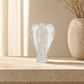 A small clear glass angel figurine with outstretched wings, displayed on a light beige stone surface next to a textured beige vase with dried stems.