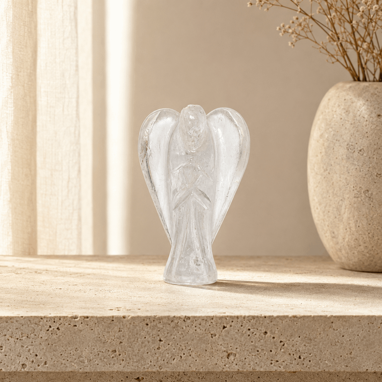 A small clear glass angel figurine with outstretched wings, displayed on a light beige stone surface next to a textured beige vase with dried stems.