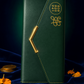 A dark green leather wallet or notebook cover with gold stitching, a gold chevron clasp, and gold monogram-style logos on the front, displayed on a dark blue velvet surface with scattered gold coins and orange petals.