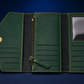 An open dark green leather wallet or organizer case with gold-tone hardware, shown on a dark blue background. The interior features card slots, a zipper compartment, elastic bands, and a clear ID window.