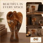 A polished brown and amber stone angel sculpture displayed on a wooden desk, with a large main view and three smaller inset scenes showing it styled on a desk, shelf, and meditation space. Text reads 'BEAUTIFUL IN EVERY SPACE' and a logo for 'Millionaire's Mind Hub' appears at the bottom.