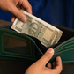 A dark green leather bifold wallet with light green contrast stitching, held open by two hands with a stack of Indian 500 Rupee notes being inserted. The wallet features a clear ID window on the front.
