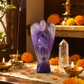 A purple amethyst angel statue stands on a wooden altar surrounded by lit brass oil lamps, a smoking incense stick holder, a clear crystal point, and orange marigold flowers.