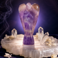 A polished purple amethyst angel figurine standing upright on a clear crystal slab, surrounded by small crystal points and dried flowers against a dark purple smoky background.