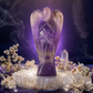 A carved amethyst angel figurine with wings and a draped figure, displayed on a white crystal base surrounded by dried flowers and crystals against a dark purple smoky background.