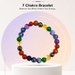 A 7 chakra bead bracelet displayed on a white cylindrical pedestal. The bracelet features colorful polished beads representing the chakras: red, orange, brown, yellow, green, blue, and purple, with one translucent amber bead.
