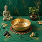 Decorative setup with a gold bowl, Buddha statue, incense, and stones on a green surface.